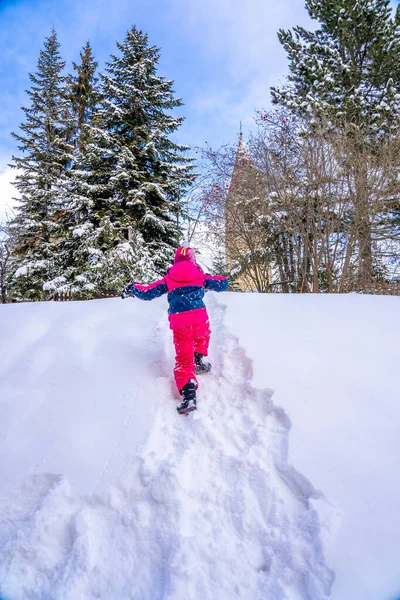 Αστείο κοριτσάκι με πολύχρωμο χειμωνιάτικο κοστούμι σκαρφαλώνει στο λόφο του χιονιού. Τα παιδιά παίζουν και πηδάνε στο χιονισμένο δάσος. Παιδί παίζει με το χιόνι το χειμώνα. — Φωτογραφία Αρχείου