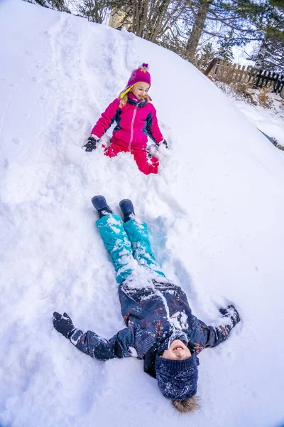 Αστείο αγόρι και κορίτσι με πολύχρωμα χειμωνιάτικα κοστούμια σκαρφαλώνει στο λόφο του χιονιού. Τα παιδιά παίζουν και πηδάνε στο χιονισμένο δάσος. Παιδί παίζει με το χιόνι το χειμώνα. — Φωτογραφία Αρχείου