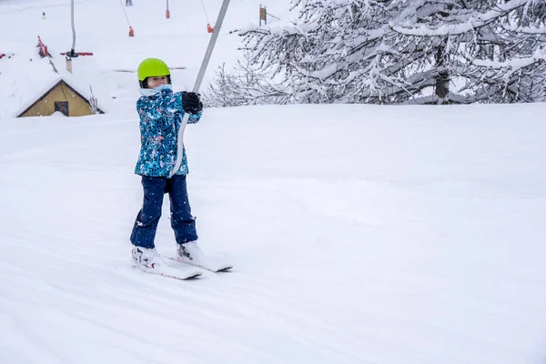 Ένα κορίτσι που σηκώνεται στο σχοινί του τελεφερίκ σκι σε μπλε φόρμα σπορ στο βουνό χιονοδρομικό κέντρο κάνει ένα μάθημα σκι κατά τη διάρκεια μιας χιονοπτώσεις. Χιονοδρομικό κέντρο στα γαλλικά βουνά. Μαλακό φόντο εστίασης — Φωτογραφία Αρχείου