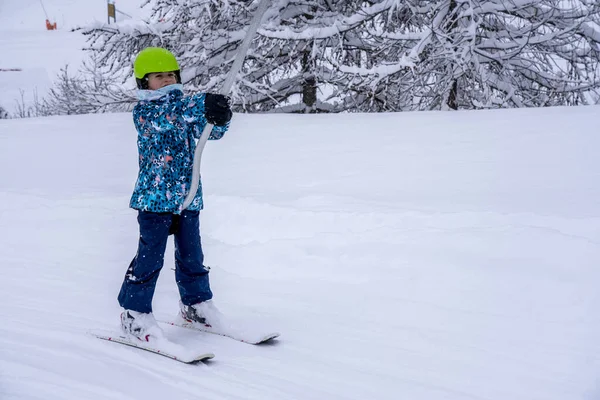Ένα κορίτσι που σηκώνεται στο σχοινί του τελεφερίκ σκι σε μπλε φόρμα σπορ στο βουνό χιονοδρομικό κέντρο κάνει ένα μάθημα σκι κατά τη διάρκεια μιας χιονοπτώσεις. Χιονοδρομικό κέντρο στα γαλλικά βουνά. Μαλακό φόντο εστίασης — Φωτογραφία Αρχείου