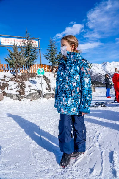 AURON, FRANCIE - 30.12.2020: Portrét dívky ubytované v horách s lékařskou maskou během koronaviru COVID-19. Dětské zimní prázdniny. Sněhová hora pozadí. — Stock fotografie