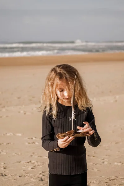 少女は手におもちゃとして木やカモメの羽の作品と砂浜に沿っています 背景にビーチの風景 高品質の写真 — ストック写真