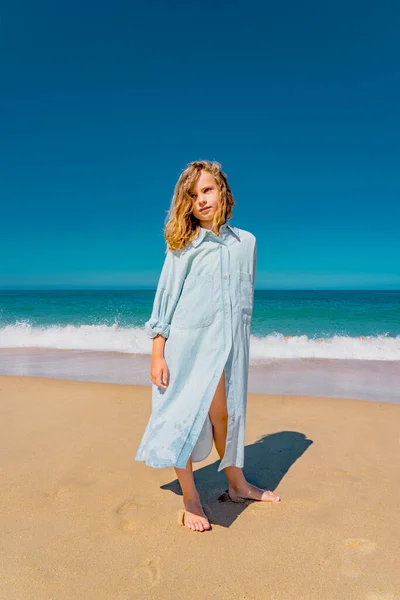Joven chica hermosa en vestido azul que permanece a lo largo de la playa de arena cerca de las olas. — Foto de Stock