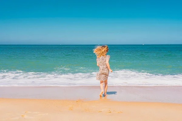 Jong mooi meisje in beige jurk lopen op het zandstrand in de buurt van de golven. — Stockfoto