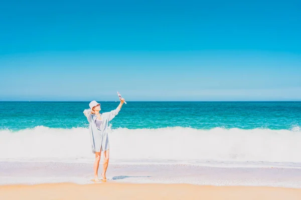 海の近くに立って自撮りしている帽子をかぶった若い女性。海と空の背景。休暇と旅行のコンセプト. — ストック写真