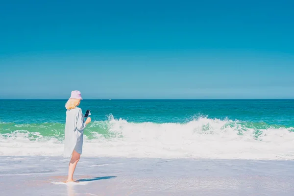 Mujer joven con sombrero de pie cerca del mar y haciendo selfie. Fondo de mar y cielo. Concepto de vacaciones y viajes. — Foto de Stock