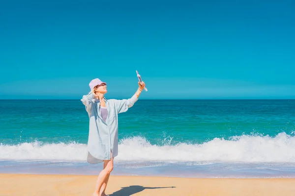 海の近くに立って自撮りしている帽子をかぶった若い女性。海と空の背景。休暇と旅行のコンセプト. — ストック写真