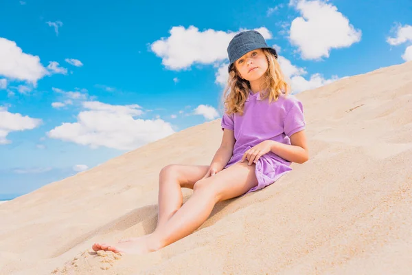 Junges nettes Mädchen mit Hut sitzt am Sandstrand. Mode-Model Mädchen posiert auf den Sanddünen. Blauer Sommerhimmel als Hintergrund. Glückliche Kindheit. — Stockfoto