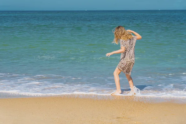 Joven chica hermosa en vestido beige corriendo en la playa de arena cerca de las olas. —  Fotos de Stock