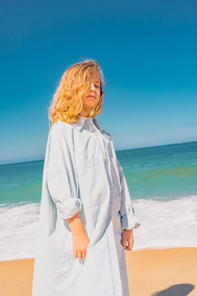Joven chica hermosa en vestido azul que permanece a lo largo de la playa de arena cerca de las olas. —  Fotos de Stock