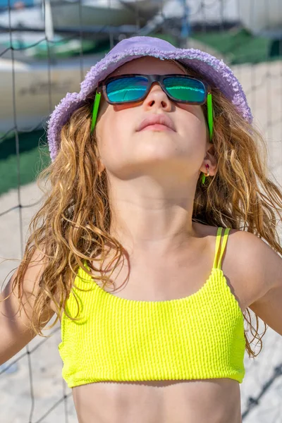 Πορτρέτο ενός όμορφου κοριτσιού με καπέλο και γυαλιά ηλίου που ποζάρει στην παραλία. Φορώντας ένα φωτεινό κίτρινο μαγιό μπικίνι. Μέρα του καλοκαιριού, λευκή άμμος. — Φωτογραφία Αρχείου