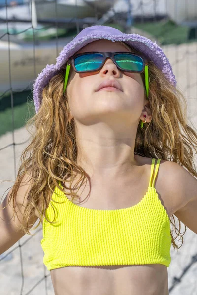 Plajda poz veren şapkalı ve güneş gözlüklü güzel bir kızın portresi. Parlak sarı bir bikini mayosu giyiyor. Yaz günü, beyaz kum. 