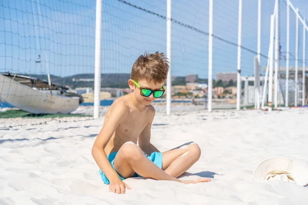 Bellissimo ragazzo in pantaloncini blu e occhiali da sole si siede sulla sabbia bianca sulla spiaggia. Gioca con sabbia bianca. — Foto Stock