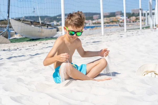 Menino bonito em calções azuis e óculos de sol se senta na areia branca na praia. Brinca com areia branca. — Fotografia de Stock