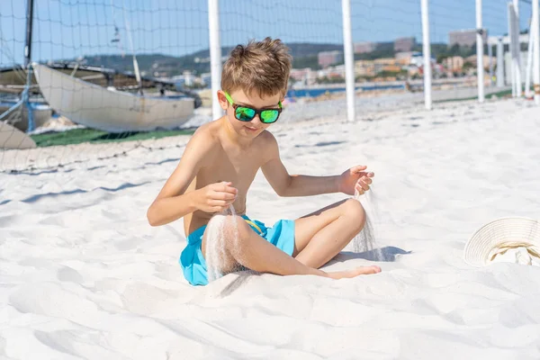 Bellissimo ragazzo in pantaloncini blu e occhiali da sole si siede sulla sabbia bianca sulla spiaggia. Gioca con sabbia bianca. — Foto Stock