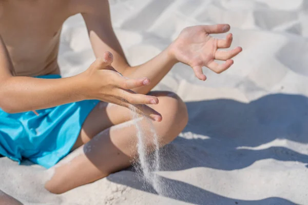 Mãos de um jovem rapaz de calções azuis senta-se na areia branca na praia. Brinca com areia branca. — Fotografia de Stock