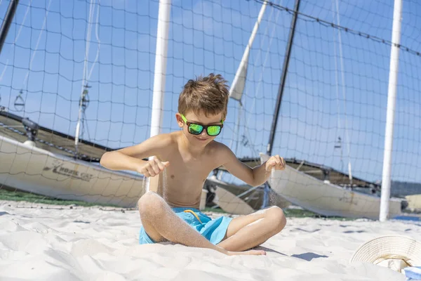 青いショートパンツとサングラスのハンサムな若い男の子は、ビーチの白い砂の上に座っています。白い砂と遊ぶ. — ストック写真