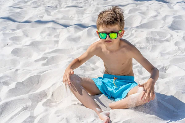 Bellissimo ragazzo in pantaloncini blu e occhiali da sole si siede sulla sabbia bianca sulla spiaggia. Gioca con sabbia bianca. — Foto Stock