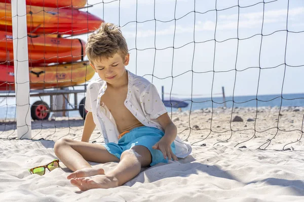 Menino bonito em calções azuis e camisa branca senta-se na areia branca na praia. Brinca com areia branca. — Fotografia de Stock