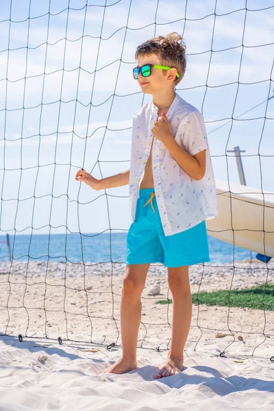 Przystojny młody chłopak w niebieskich spodenkach i białej koszuli, w okularach przeciwsłonecznych stojący na białym piasku na boisku do siatkówki plażowej. — Zdjęcie stockowe
