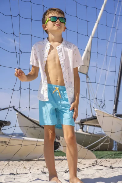 Stilig ung pojke i blå shorts och vit skjorta, klädd i solglasögon stående på vit sand på en volleyboll strandbana. — Stockfoto