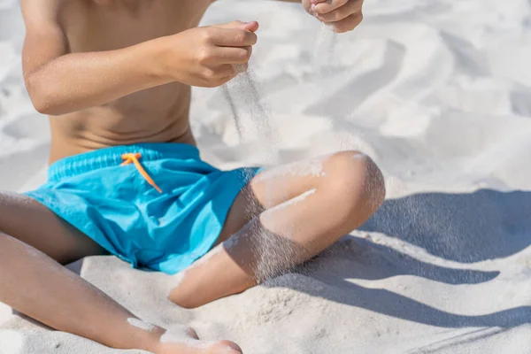 Ręce małego chłopca w niebieskich spodenkach siedzą na białym piasku na plaży. Zabawa z białym piaskiem. — Zdjęcie stockowe