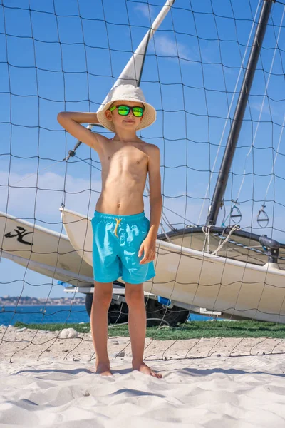 Beau jeune garçon en short bleu, lunettes de soleil et chapeau debout sur sable blanc sur un terrain de volley-ball de plage. — Photo