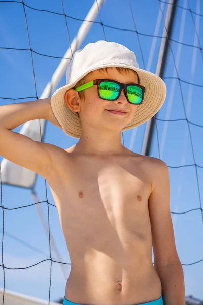 Beau jeune garçon en short bleu, lunettes de soleil et chapeau debout sur sable blanc sur un terrain de volley-ball de plage. — Photo