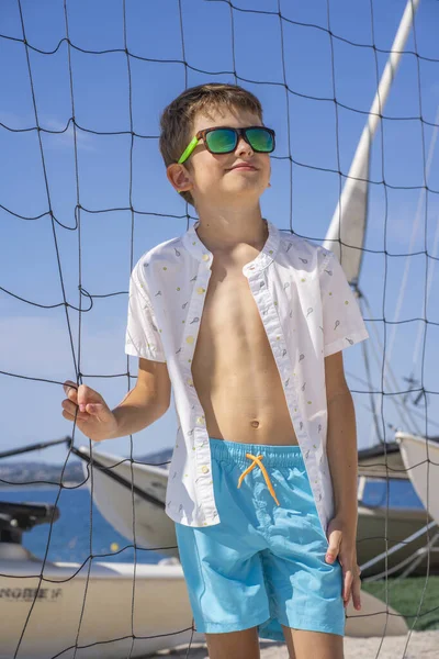 Beau garçon en short bleu et chemise blanche, portant des lunettes de soleil debout sur du sable blanc sur un terrain de volley-ball de plage. — Photo