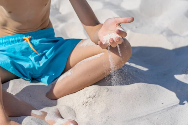 Mãos de um jovem rapaz de calções azuis senta-se na areia branca na praia. Brinca com areia branca. — Fotografia de Stock