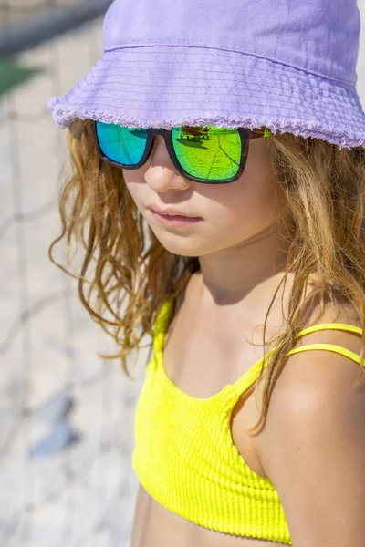 Portret młodej, pięknej modelki w kapeluszu i okularach przeciwsłonecznych na plaży. W jasnożółtym bikini. Letni dzień, biały piasek. — Zdjęcie stockowe