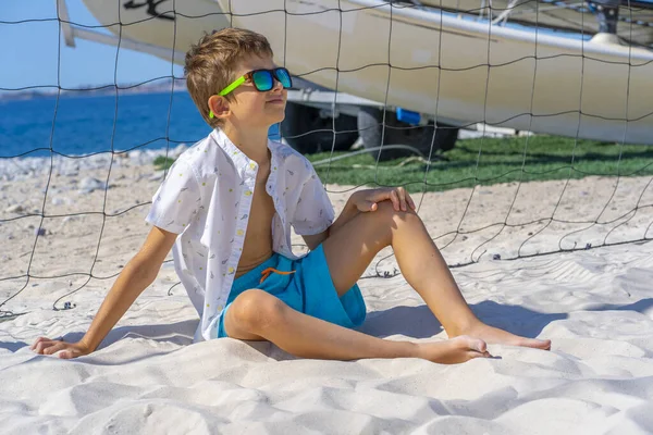 Bellissimo ragazzo in pantaloncini blu, occhiali da sole e camicia bianca si siede sulla sabbia bianca sulla spiaggia. Gioca con sabbia bianca. — Foto Stock