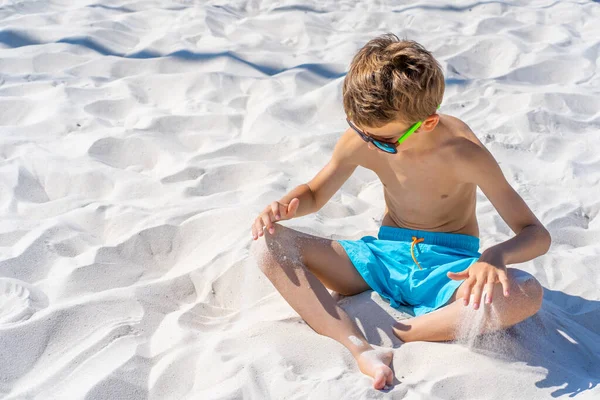 Bellissimo ragazzo in pantaloncini blu e occhiali da sole si siede sulla sabbia bianca sulla spiaggia. Gioca con sabbia bianca. — Foto Stock