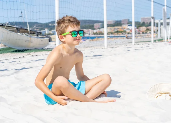 青いショートパンツとサングラスのハンサムな若い男の子は、ビーチの白い砂の上に座っています。白い砂と遊ぶ. — ストック写真