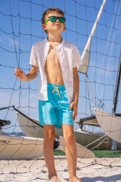 Menino bonito em shorts azuis e camisa branca, usando óculos de sol em pé na areia branca em uma quadra de praia de voleibol. — Fotografia de Stock