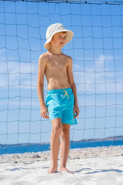 Przystojny młody chłopiec w niebieskich spodenkach i kapeluszu stojący na białym piasku na boisku do siatkówki plażowej. — Zdjęcie stockowe