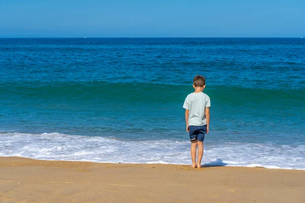夏のビーチの波の近くに一人の少年が立っている。若い10代の若者は、後ろから海を見ています。夏休み旅行のコンセプト.. — ストック写真
