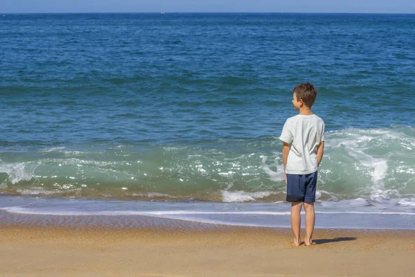 Um menino fica perto das ondas na praia de verão. Jovem adolescente, vista pelas costas, olha para o oceano. Verão férias férias viagem conceito.. — Fotografia de Stock