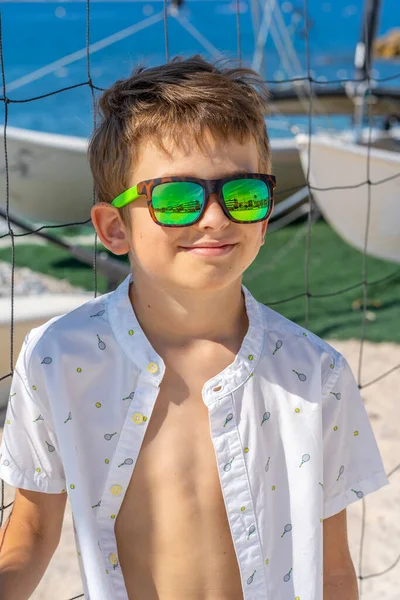 Menino bonito em shorts azuis e camisa branca, usando óculos de sol em pé na areia branca em uma quadra de praia de voleibol. — Fotografia de Stock