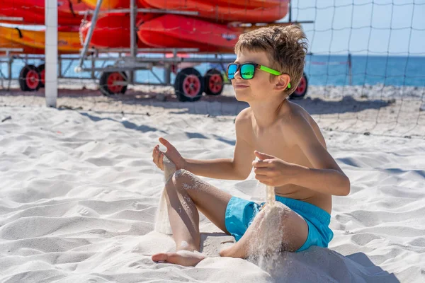 Bellissimo ragazzo in pantaloncini blu e occhiali da sole si siede sulla sabbia bianca sulla spiaggia. Gioca con sabbia bianca. — Foto Stock