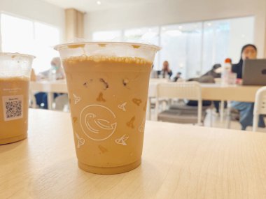 Açık renkli ahşap bir masada otururken soğuk, buzlu bir sütlü kahve ya da temiz bir plastik bardakta latte yakın çekim..