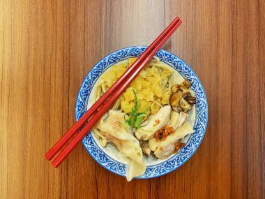 Resim, sıcak, yatay tahıllı ahşap bir masa üzerine kurulmuş mavi ve beyaz bir seramik kasede servis edilen erişte kâsesini yukarıdan aşağıya doğru gösteriyor..