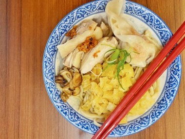 Resim, sıcak, yatay tahıllı ahşap bir masa üzerine kurulmuş mavi ve beyaz bir seramik kasede servis edilen erişte kâsesini yukarıdan aşağıya doğru gösteriyor..