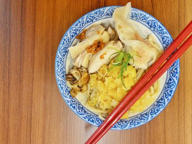 Resim, sıcak, yatay tahıllı ahşap bir masa üzerine kurulmuş mavi ve beyaz bir seramik kasede servis edilen erişte kâsesini yukarıdan aşağıya doğru gösteriyor..