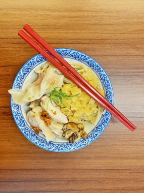 Resim, sıcak, yatay tahıllı ahşap bir masa üzerine kurulmuş mavi ve beyaz bir seramik kasede servis edilen erişte kâsesini yukarıdan aşağıya doğru gösteriyor..