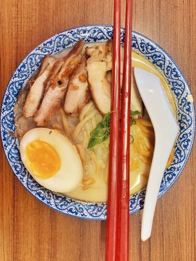 Ilık ahşap bir masanın üstünde duran mavi ve beyaz desenli seramik kasede titizlikle düzenlenmiş klasik bir ramen kasesinin canlı, tepeden tırnağa görüntüsü..