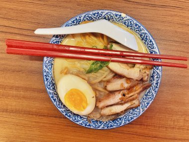 Ilık ahşap bir masanın üstünde duran mavi ve beyaz desenli seramik kasede titizlikle düzenlenmiş klasik bir ramen kasesinin canlı, tepeden tırnağa görüntüsü..