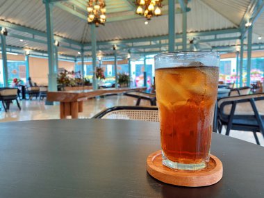 Koyu, yuvarlak bir masada ön planda belirgin bir şekilde duran ferahlatıcı bir bardak buzlu çay. Çay önü açık bir bardakta sunuluyor. Tahta bir bardakta dinleniyor. Üstünde görünen buz küpleri yüzüyor..