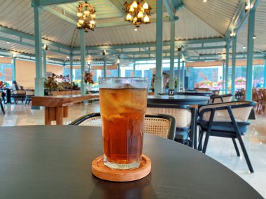 Koyu, yuvarlak bir masada ön planda belirgin bir şekilde duran ferahlatıcı bir bardak buzlu çay. Çay önü açık bir bardakta sunuluyor. Tahta bir bardakta dinleniyor. Üstünde görünen buz küpleri yüzüyor..