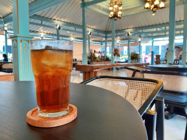 Koyu, yuvarlak bir masada ön planda belirgin bir şekilde duran ferahlatıcı bir bardak buzlu çay. Çay önü açık bir bardakta sunuluyor. Tahta bir bardakta dinleniyor. Üstünde görünen buz küpleri yüzüyor..